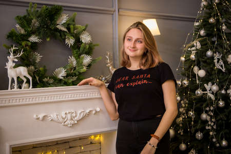 a young blonde girl in a New Year's t-shirt stands by the tree near the fireplace in New Year's decorationsの写真素材