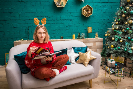 young blonde girl in a hoop with horns in a red New Year's sweater sits on the sofa and reads a book in the New Year's decorationsの写真素材