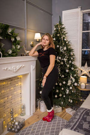 a young blonde girl in a New Year's t-shirt stands by the tree near the fireplace in New Year's decorationsの写真素材