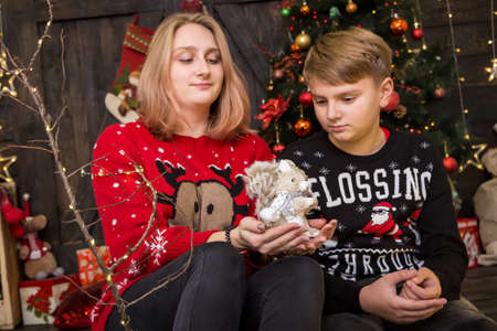 handsome boy in a New Year's sweater sits with a girl and a toy squirrel on the sofa near a decorated Christmas treeの写真素材