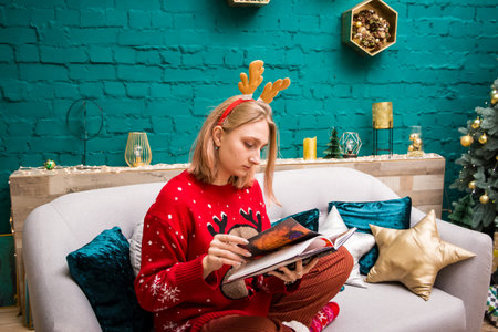 young blonde girl in a hoop with horns in a red New Year's sweater sits on the sofa and reads a book in the New Year's decorationsの写真素材