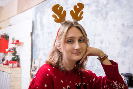 young blonde girl in a hoop with horns in a red New Year's sweater stands in a photo studio of New Year's decorationsの写真素材