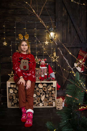 a young blonde girl in a hoop with horns sitting in a red New Year's sweater by a woodpile in New Year's decorationsの写真素材