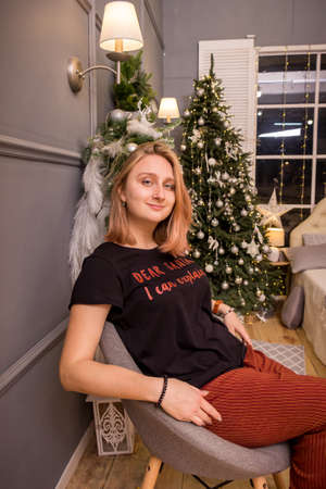young blonde girl in a new year t-shirt sits in a chair by the tree near the fireplace in New Year's decorationsの写真素材