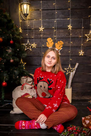 young blonde girl in a hoop with horns sitting in a red New Year's sweater with crockery in New Year's decorationsの写真素材