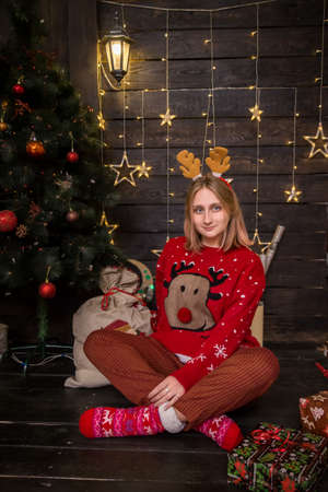 young blonde girl in a hoop with horns sitting in a red New Year's sweater with crockery in New Year's decorationsの写真素材