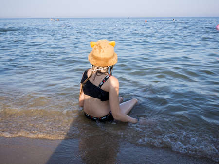a young girl in a hat with ears against the backdrop of the blue sea in the water with her back to the lens near the seashoreの写真素材