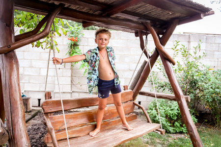 the boy stands on a wooden swing in a colorful shirt and shorts, barefootの写真素材