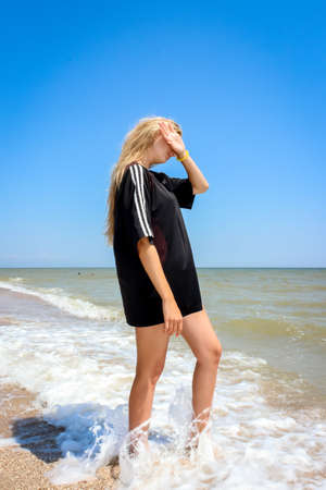 young girl schoolgirl in a big black T-shirt on the azure seashore on a sandy beachの写真素材