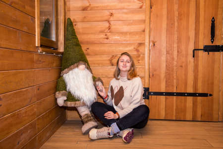 blonde girl with a forest gnome sits in a winter, log houseの写真素材
