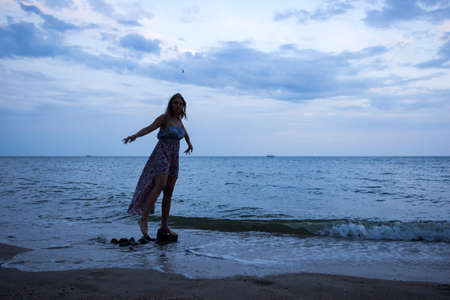 a young girl in the evening on the seashore in a sundress and with a hat, sunset, blue sky, clouds, calm, surf, sand, empty beach, wildの写真素材