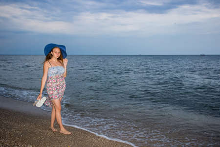 a young girl in the evening on the seashore in a sundress and with a hat, sunset, blue sky, clouds, calm, surf, sand, empty beach, wildの写真素材