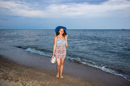 a young girl in the evening on the seashore in a sundress and with a hat, sunset, blue sky, clouds, calm, surf, sand, empty beach, wildの写真素材