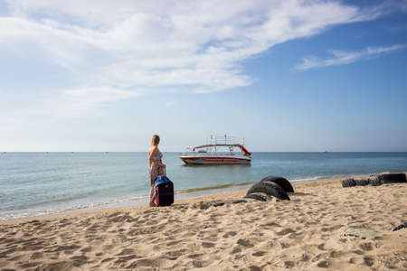 a young girl in a sundress and with a hat stands with a suitcase on the beach near the boat near the seaの写真素材