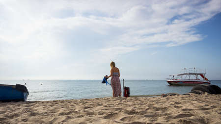 a young girl in a sundress and with a hat stands with a suitcase at the water's edge on the beachの写真素材
