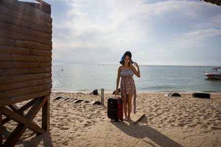a young girl stands with a suitcase on a sandy seashore in a sundress and with a hatの写真素材