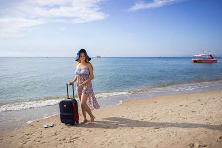 a young girl stands with a suitcase on a sandy seashore in a sundress and with a hatの写真素材