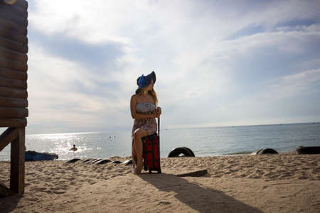 a young girl sits on a suitcase on a sandy seashore in a sundress and with a hatの写真素材