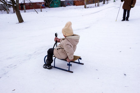 little girl rides down  slide on  sled, in a snowy park, rear view, lollipops, family, full length, children ride, walk, winter, snowy, winter day, rear view,の写真素材