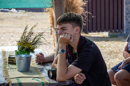 dark-haired teenager boy sits at table in street wooden gazebo, with camera, photographer, propping...