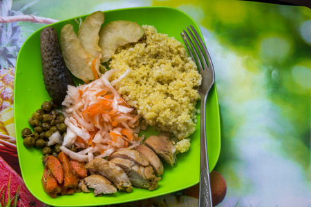 green plate with yellow cereal, sliced chicken, sauerkraut salad, vegetables, green peas, stewed carrots, soaked apples, cucumber, millet, couscous, fork in plate, healthy eating,の写真素材