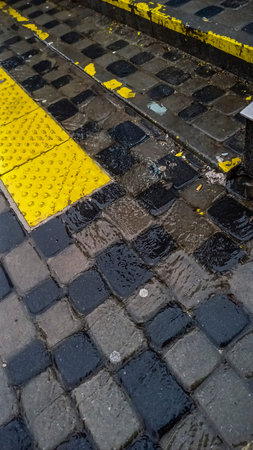 water flows on city street on gray cobblestones, tactile tiles of bright yellow color, marking,...
