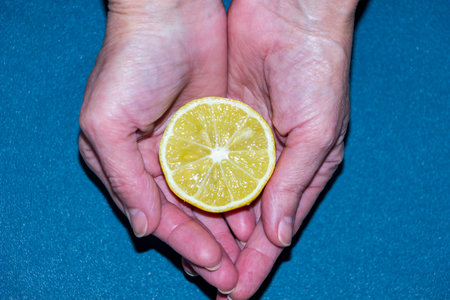 yellow half of lemon in woman's palms, hands holding citrus, useful, daylight, close-up, food, vitamins, blue backgroundの写真素材