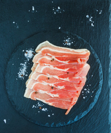 Raw sliced bacon on black slate plate, top view. Toned.の写真素材