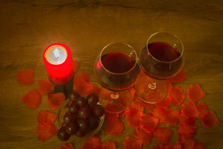 Red wine glasses, grapes, burning candle, and rose leaves on tableの写真素材