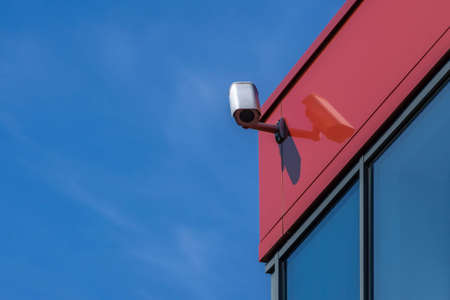 CCTV surveillance camera installed on modern red building wall. Security camera on blue sky...