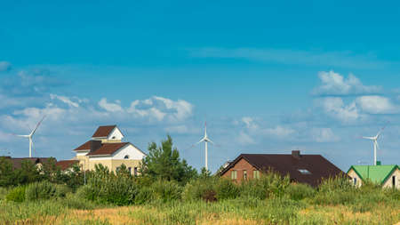 Wind turbines behind houses in residential area on blue sky background. Friendly environment, clean and renewable energy conceptの写真素材