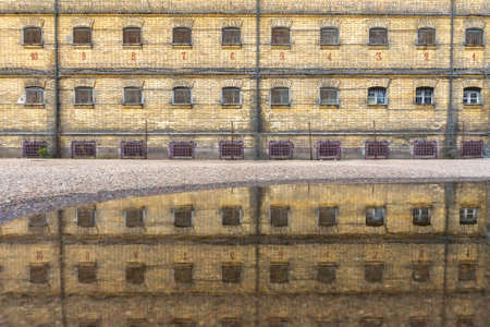 Old abandoned prison brick wall and cell windows with metal bars, jail wall reflection in water on ground after rainの写真素材