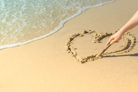 Hand of woman drawing heart on sand in beach near sea wave. Copy spaceの写真素材