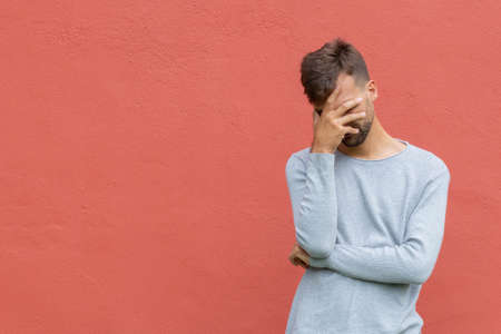 Facepalm. Ashamed embarrassed man covering his face. Young guy on red wall background with copy space. Emotion facial expression and feelings conceptの写真素材