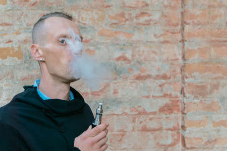 Man vaping electronic cigarette or vape outdoor on vintage brick wall background with copy space. Guy smoke vaporizer. Vapor and alternative nicotine free smoking concept.の写真素材