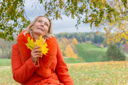 Pretty middle-aged woman in orange coat holding autumn maple leaves outside in colorful fall nature background. Copy space in right side. Portrait of romantic femaleの写真素材