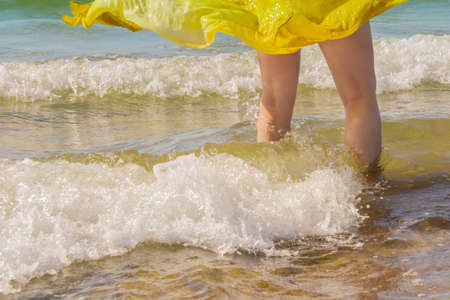 Female legs covered yellow scarf floating in wind on sandy coast in wavy sea. Scarf on standing...