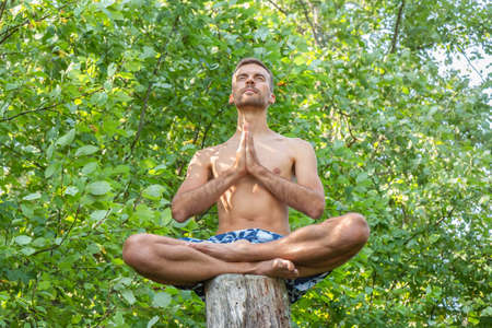 Young man with his hands together sitting on cut off old tree trunk outdoors in forest. Man meditating in lotus pose on tree stem. Guy man feeling free in forest. Peace and relaxation concept.の写真素材