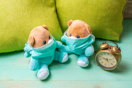 Two teddy bears with protective masks leaning against the pillow and alarm clock at home. Medicine masks on bear toys and clock. Stay at home and be safe conceptionの写真素材