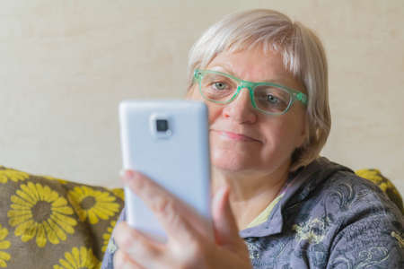 Elderly woman using mobile phone for connection with relatives. Senior woman taking selfie. Lady with smartphone in hand at home. Mature female using cellphone. Quarantine during coronavirus pandemic.の写真素材
