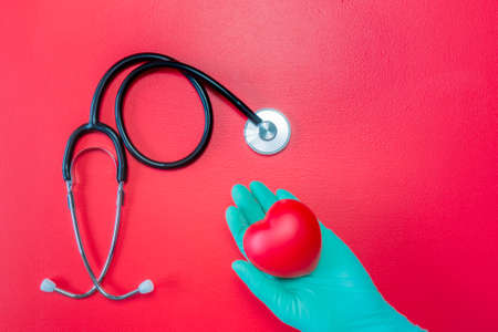 Medical stethoscope and hand in green rubber glove holding red heart on red background. Flat lay of isolated of heart in hand in glove and stethoscope. Mockup for organ donation concept. Copy space.の写真素材