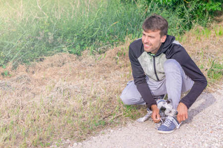 Young man getting ready to run outdoors on sunny day. Puppy helping to tie jogging shoes of guy....