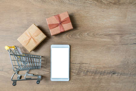 Smartphone and gift boxes flying to shopping cart on wood background. Shopping trolley, boxes and phone on table. Top view, copy space. Showing fast order delivery. Mobile shopping on internet conceptの写真素材