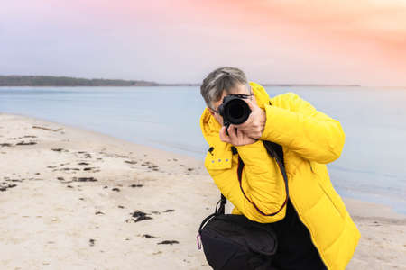 Male photographer taking photos on beach. Traveler photographer in yellow jacket looking straight through camera on sea coast. Travel photography of sea landscape on vacation.の写真素材