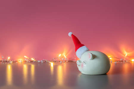 Piggy bank in the hat of Santa Claus and shining garland on pink background. Concept of savings for Christmas and New Year, holiday. Copy space for text, banner. Christmas time.の写真素材