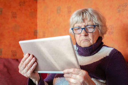 Senior woman in eyeglasses using tablet and sitting on couch at home. Grandmother holding digital...