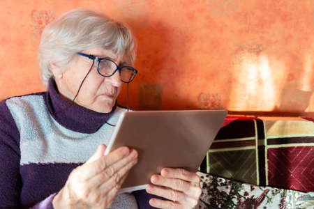 Senior woman in eyeglasses looking tablet and sitting on couch at home. Old lady looking at tablet. Gray hair grandmother holding digital device in hands. Gray hair Modern pensioner lifestyle concept.の写真素材