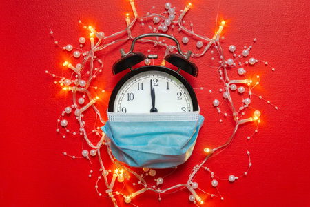 Garland lights around alarm clock covering medical mask showing midnight. Clock with protective mask at New Year's night on red background. Safe Christmas and New Year concept.の写真素材