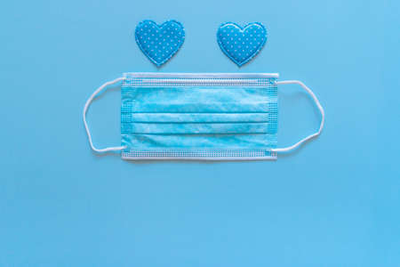 Medical protective face mask and two blue heart shapes as eyes on blue background. Disposable...