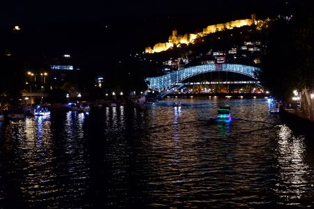 Tbilisi, Georgia - September 2019: The Bridge of Peace is a bow-shaped pedestrian bridge, a steal and glass construction illuminated with numerous LEDs, over the Kura River in downtown Tbilisi, capital of Georgia. Night photo.の写真素材